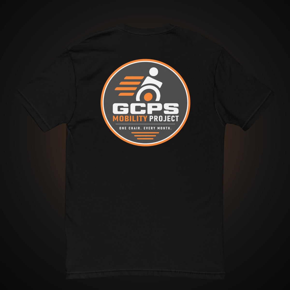 Back of the black GCPS Mobility Project T-Shirt with a large orange, gray and white GCPS Mobility Project badge reading "One Chair. Every Month."